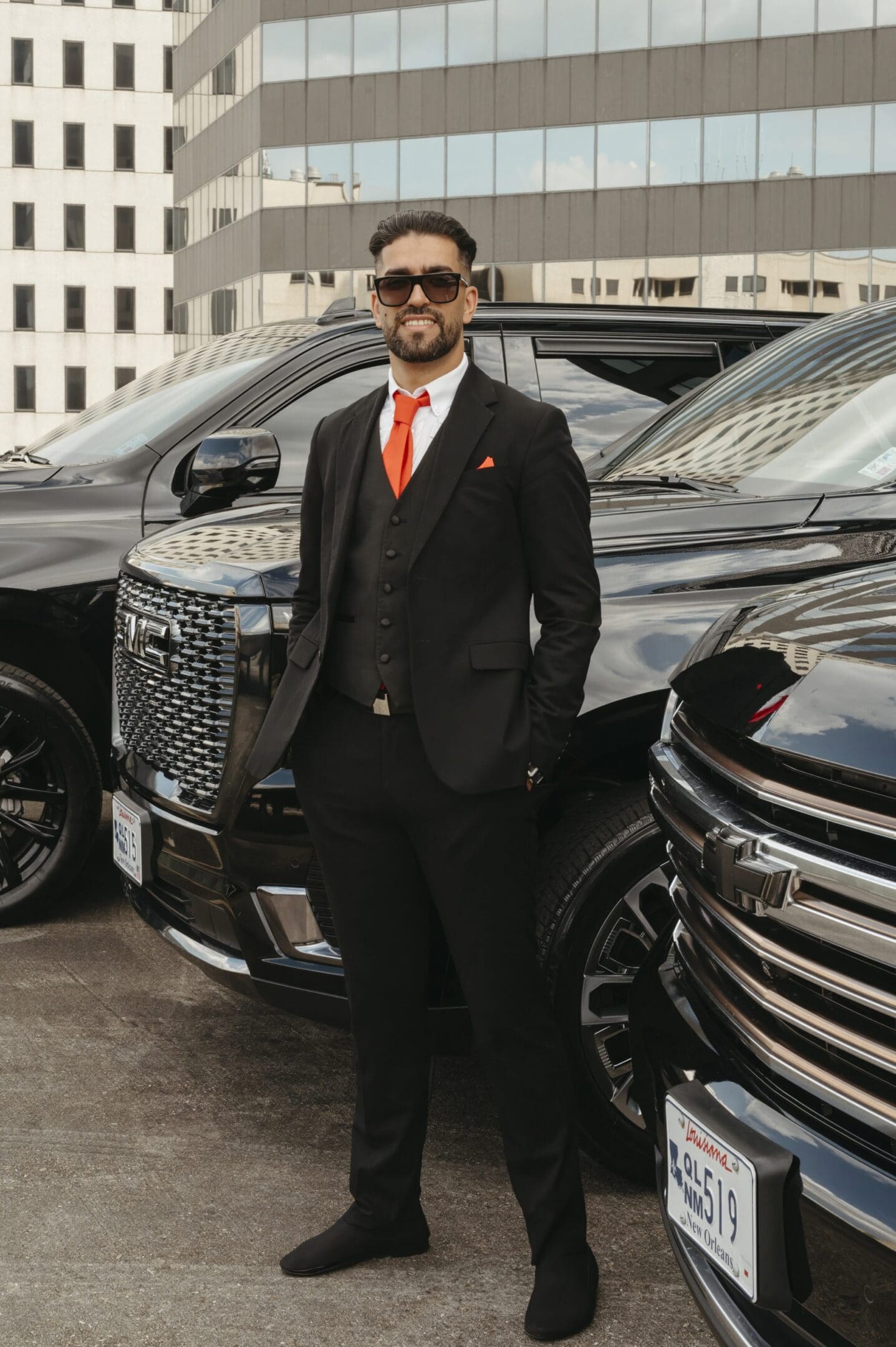 A man in suit and tie standing next to two black trucks.